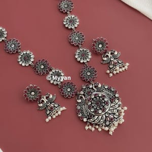 May include: A silver necklace with a large pendant featuring a peacock design and pink stones. The necklace is adorned with smaller circular pendants with pink stones. Two matching earrings with pink stones and a peacock design are also included.