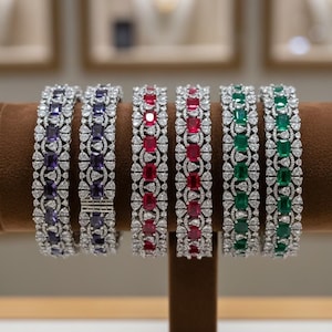May include: Six gemstone bracelets on display. The bracelets feature alternating rectangular and round-cut gemstones in purple, red, and green, set in a silver filigree design. The bracelets are displayed on a brown stand.