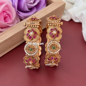 May include: A pair of gold bangles with intricate designs featuring pink and green gemstones. The bangles are decorated with white pearls and have a floral motif.