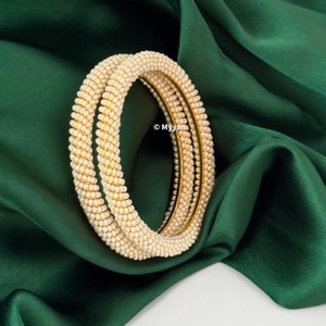 May include: A set of two white pearl bangles with a gold base. The bangles are stacked on top of each other and are lying on a green fabric background.