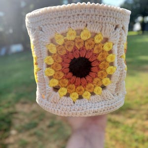 May include: A cream-coloured crocheted cosy with a sunflower design. The sunflower is in shades of yellow, orange, and brown. The crochet work is visible, highlighting the texture and craftsmanship of the handmade item. A decorative item.