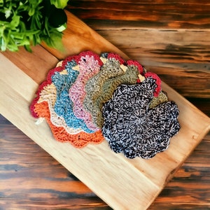 May include: A collection of handmade crochet coasters in various colors, including orange, blue, pink, and black and white. Each coaster has a scalloped edge and is arranged on a wooden cutting board.