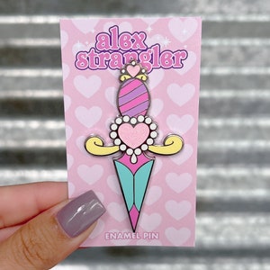 May include: Pink and blue enamel pin with a heart-shaped dagger design. The pin has a yellow handle and a crown on top. The text "alex strangler" and "enamel pin" are on the card.