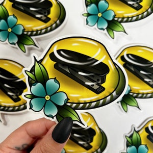 May include: A yellow jelly-like sticker with a black stapler inside and a blue flower with green leaves on the side.