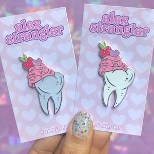May include: Two enamel pins featuring a tooth with a pink and white frosting and strawberry topping. The pins are on pink cards with white hearts and the text "alex strangler" in pink letters.