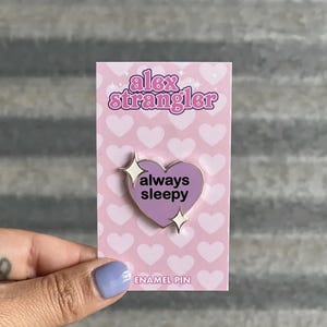 May include: A lavender heart-shaped enamel pin with the words "always sleepy" in white text. The pin is on a pink card with a heart pattern and the text "alex strangler". Two starbursts flank the heart.
