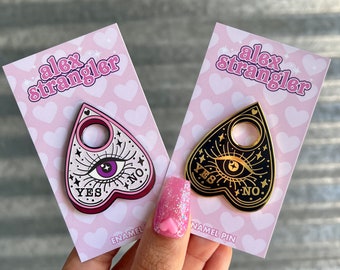 Puede incluir: Dos pines de esmalte en forma de tablero de Ouija en forma de corazón con un solo ojo y las palabras "YES" y "NO" en cada uno. Los pines están en tarjetas rosas con corazones blancos y el texto "alex strangler" en cada tarjeta.