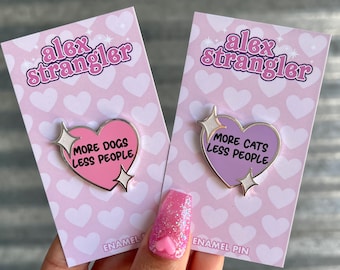 May include: Two enamel pins in the shape of hearts with the text "More Dogs Less People" and "More Cats Less People". The pins are on pink and purple backgrounds with a heart pattern.