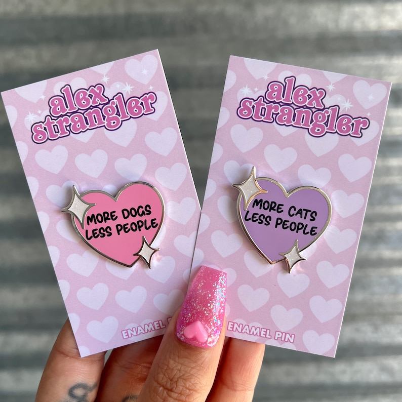May include: Two enamel pins in the shape of hearts with the text "More Dogs Less People" and "More Cats Less People". The pins are on pink and purple backgrounds with a heart pattern.