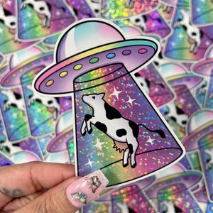 May include: A holographic sticker featuring a black and white cow being abducted by a rainbow-colored UFO. The sticker has a glittery finish.