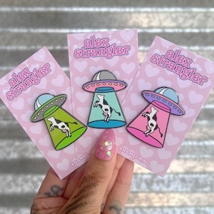 May include: Three enamel pins featuring a cow in a UFO. The pins are on pink cards with the text "alex strangler" and "enamel pin".