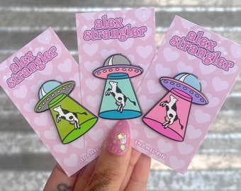 May include: Three enamel pins featuring a cow in a UFO. The pins are on pink cards with the text "alex strangler" and "enamel pin".