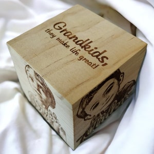 May include: A light-colored wooden cube with engraved text that reads "Grandkids, they make life great!" The cube also features engraved images of children. The cube is resting on a white surface.