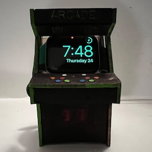 May include: A wooden arcade-style stand with a green trim, designed to hold a smartwatch. The stand has a black background with a red and white "11" on the front. The smartwatch is displaying the time "7:48" and the date "Thursday 24".