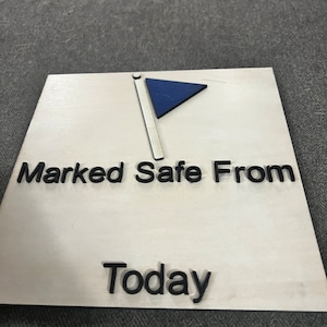 May include: White wooden sign with black text that reads "Marked Safe From Today". A blue triangle flag with a white pole is in the top left corner.