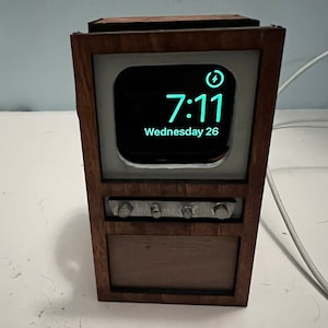 Può includere: Una replica in legno di una televisione vintage con uno schermo bianco che mostra un orologio digitale che segna le 7:11 e la data mercoledì 26. L'orologio è in carica con un'icona di fulmine.
