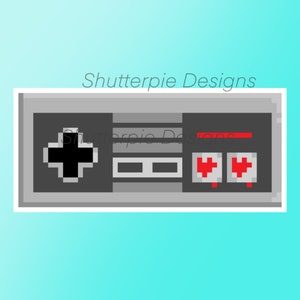 Puede incluir: Una ilustración pixelada de un mando de videojuegos gris clásico con un joystick negro, dos botones rojos y el texto "Shutterpie Designs" en la parte superior.