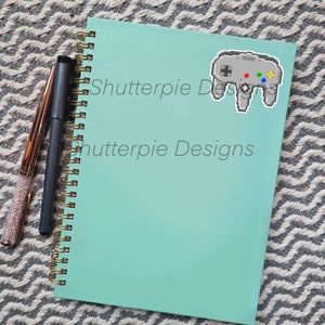 May include: A teal notebook with a gold spiral binding. The notebook has the text "Shutterpie Designs" printed on the front cover. A pixelated image of a grey Nintendo 64 controller is on the front cover.
