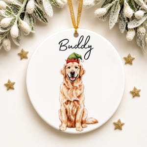 May include: A round white ceramic ornament featuring a golden retriever illustration wearing a green and red elf hat. The name Buddy is printed in black script above the dog. A gold ribbon is attached to the top for hanging purposes.