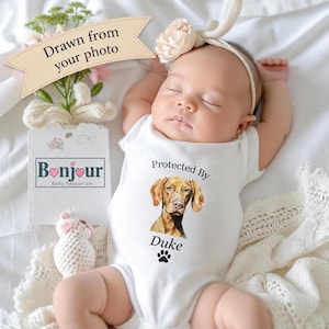 May include: A white baby bodysuit featuring a watercolour illustration of a brown dog, the text "Protected By Duke," and a black paw print. A tag displays "Drawn from your photo" and "Bonjour Baby Emporium" with a small floral graphic.