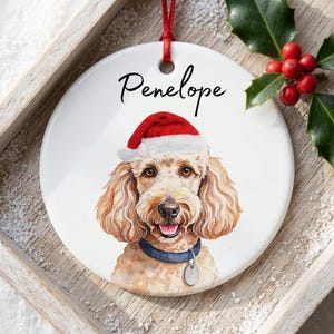 Può includere: Un ornamento rotondo in ceramica bianca con un'illustrazione ad acquerello di un cane marrone chiaro dal pelo riccio che indossa un cappello da Babbo Natale rosso e bianco e un collare blu con una medaglietta argentata. Il nome Penelope è stampato.