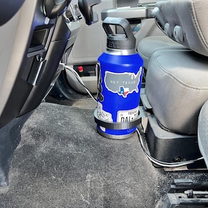 May include: A blue insulated water bottle with a black handle and stickers, including one that says "NOT TEXAS." The bottle is in a black cup holder in a vehicle. A white and black cable is visible.