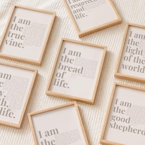 May include: A collection of six rectangular wall art prints in light wood frames. Each print features grey text on a white background, displaying phrases such as I am the true vine, I am the bread of life, I am the light of the world, and I am the way, the truth, and life.