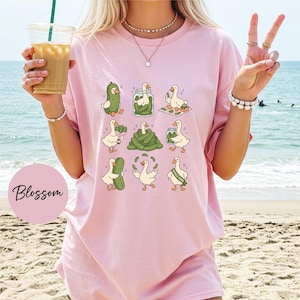 A woman wearing a pink t-shirt with a graphic of cartoon ducks holding pickles, standing on a beach with the ocean in the background.