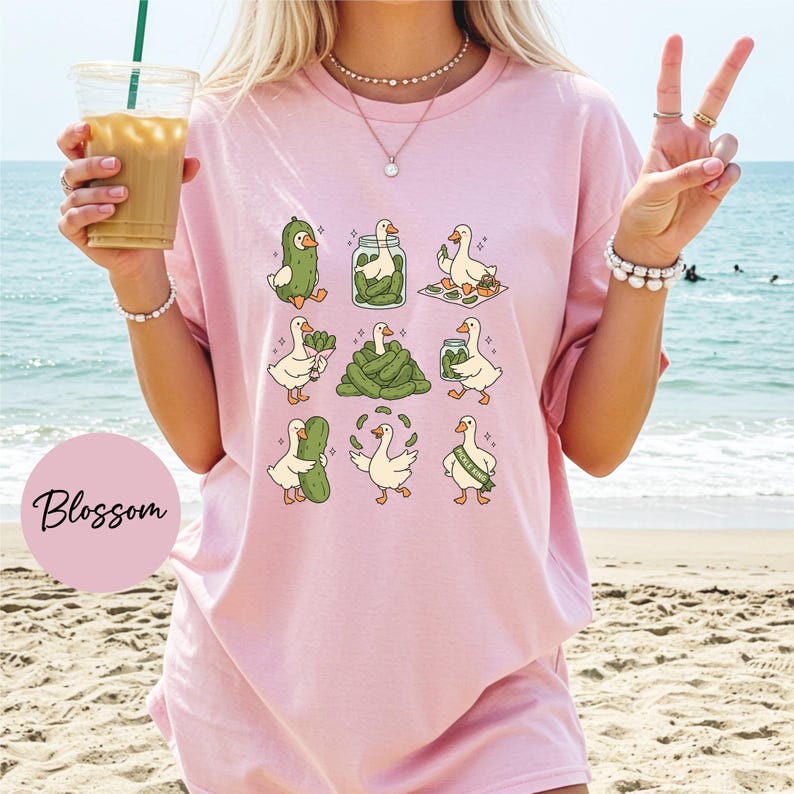 A woman wearing a pink t-shirt with a graphic of cartoon ducks holding pickles, standing on a beach with the ocean in the background.