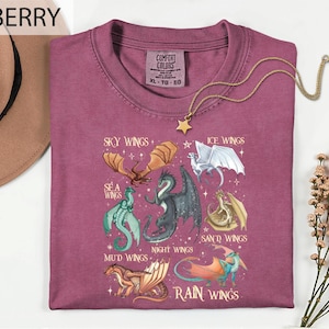 May include: Berry-coloured t-shirt featuring a graphic of different dragon wings, such as Sky, Ice, Sea, Mud, Night, Sand, and Rain Wings. A gold star pendant necklace is draped over the shirt. The shirt is labelled XL-TO-EG.