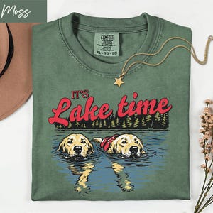 Könnte beinhalten: Ein moosgrünes T-Shirt mit dem Schriftzug "It's Lake Time" in roter Schrift. Die Grafik zeigt zwei Golden Retriever, die in einem See schwimmen. Eine goldene Sternanhänger-Halskette liegt auf dem Shirt. Das Shirt ist ein Comfort Colors XL.