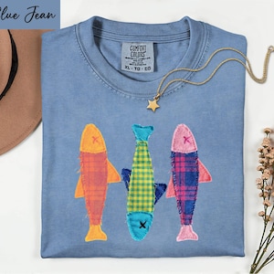 May include: A blue jean colored t-shirt with a Comfort Colors label at the neck. The shirt features three appliqued fish designs in orange, yellow-green, and pink plaid patterns, each with a stitched X for an eye. A gold star pendant necklace is included.