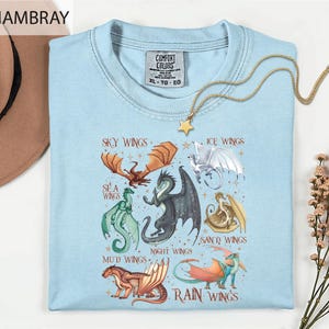 A light blue t-shirt with a graphic design of various dragon-like creatures, including sea, night, mud, sand, rain, and ice wings, along with the text "Sky Wings", "Ice Wings", "Sea Wings", "Night Wings", "Mud Wings", and "Sand Wings".