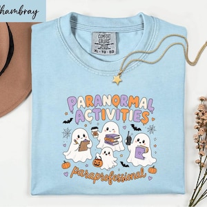May include: A light blue t-shirt with a graphic print featuring four white ghosts, pumpkins, bats, and spiderwebs. The text reads "PARANORMAL ACTIVITIES" and "paraprofessional". The shirt has a Comfort Colors label in size XL and a gold star necklace.