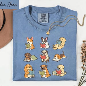 May include: A blue t-shirt featuring a graphic print of nine different dog breeds, each reading a book. The shirt has a Comfort Colors brand label at the neck, indicating size XL. A gold-toned necklace with a star charm rests on the fabric.