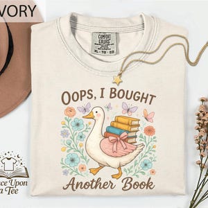 May include: Ivory-colored t-shirt with the text "Oops, I Bought Another Book." The graphic features a cartoon duck carrying books, surrounded by flowers and butterflies. A gold necklace with a star pendant is draped on the shirt.