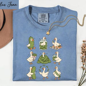A blue t-shirt with a graphic of nine cartoon ducks, each performing a different action, such as eating pickles, standing on one leg, or holding a jar of pickles. The t-shirt is laid out on a white surface, with a hat and some flowers visible in the background.
