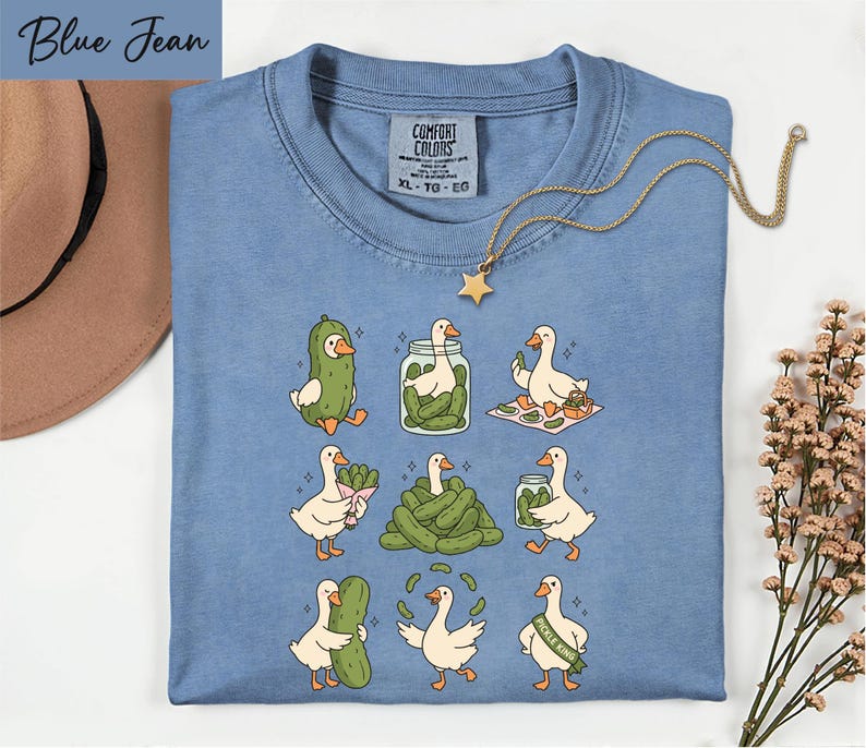 A blue t-shirt with a graphic of nine cartoon ducks, each performing a different action, such as eating pickles, standing on one leg, or holding a jar of pickles. The t-shirt is laid out on a white surface, with a hat and some flowers visible in the background.