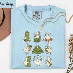 A light blue t-shirt with a cartoon illustration of nine cartoon ducks, each performing a different action, such as eating pickles, standing on one leg, or walking on a hopscotch pattern. The t-shirt is displayed on a white surface, with a hat and some flowers in the background.