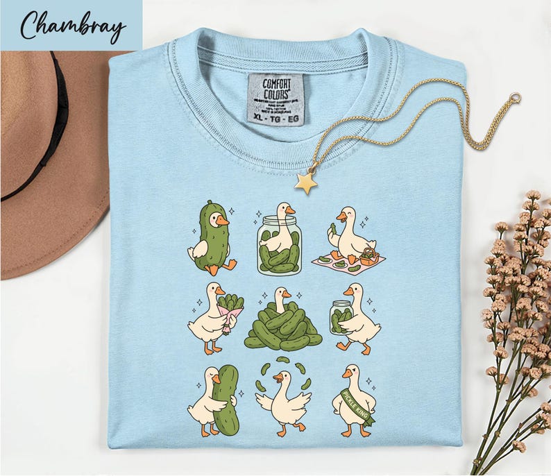 A light blue t-shirt with a cartoon illustration of nine cartoon ducks, each performing a different action, such as eating pickles, standing on one leg, or walking on a hopscotch pattern. The t-shirt is displayed on a white surface, with a hat and some flowers in the background.