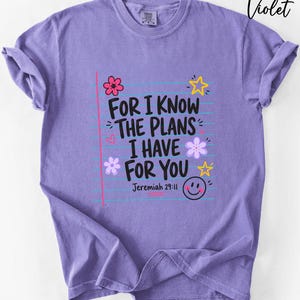 May include: A violet short-sleeve t-shirt featuring a graphic print of notebook paper lines with the black text "FOR I KNOW THE PLANS I HAVE FOR YOU" and "Jeremiah 29:11". The design includes colorful doodles of flowers, stars, a heart, and a smiley face.