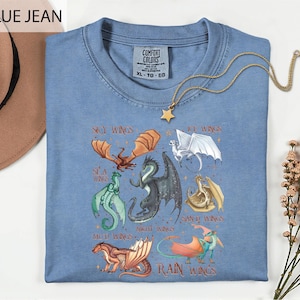 A blue t-shirt with a graphic design of various dragon characters, including a red dragon, a green dragon, and a white dragon, along with the text "SKY WINGS" and "ICE WINGS". The t-shirt is displayed on a white surface, with a hat and some flowers in the background.