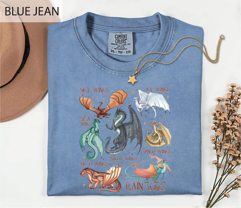 A blue t-shirt with a graphic design of various dragon characters, including a red dragon, a green dragon, and a white dragon, along with the text "SKY WINGS" and "ICE WINGS". The t-shirt is displayed on a white surface, with a hat and some flowers in the background.
