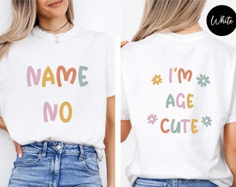 May include: A white short-sleeve t-shirt featuring colorful text. The front displays the words NAME NO in pastel tones. The back features the text I'M AGE CUTE arranged with small flower graphics in yellow, green, and pink, all in a rounded font.