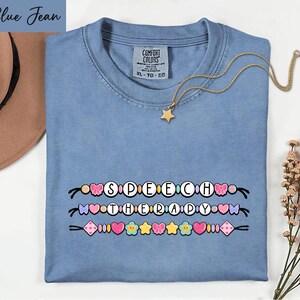 May include: A blue jean colored t-shirt with a graphic print of three rows of colorful beads spelling out SPEECH THERAPY. The beads include hearts, stars, flowers, and bows. A gold chain necklace with a small star charm rests on the shirt.
