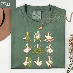 Pode incluir: Uma camiseta Comfort Colors verde musgo com um design lúdico de patos de desenho animado e picles. Os patos são retratados com picles, em potes e como o "Pickle King". Um colar com pingente de estrela dourada está sobre a camisa.