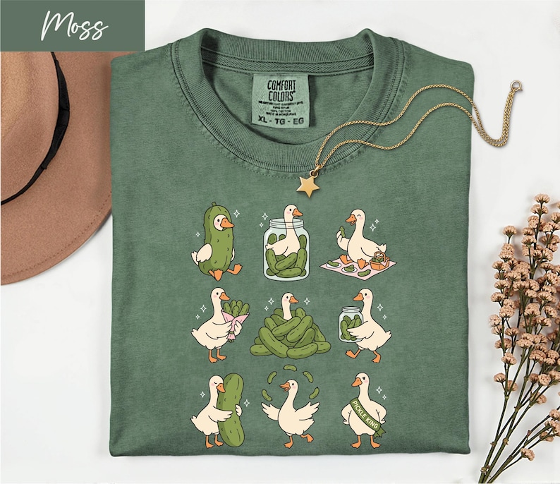 a green t-shirt with a cartoon illustration of nine cartoon ducks, each performing a different action, such as eating pickles, standing on one leg, or dancing. The t-shirt is laid out on a white surface, with a brown hat and some flowers visible in the background.