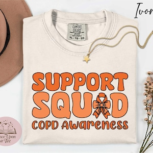 Puede incluir: Camiseta de color marfil con la frase "SUPPORT SQUAD COPD Awareness" en naranja. También se ve un collar dorado con un colgante de estrella y un sombrero marrón.