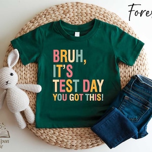 May include: A forest green t-shirt with the text "BRUH, IT'S TEST DAY YOU GOT THIS!" in colorful block letters. The shirt is paired with blue jeans and a stuffed bunny on a woven mat. The words "Once Upon a Tee" are at the bottom.