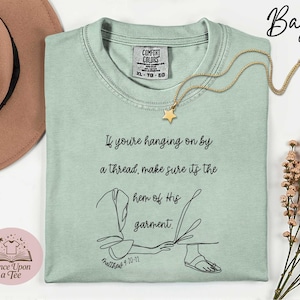 May include: A pale green Comfort Colors t-shirt with the text "If you're hanging on by a thread, make sure its the hem of His garment." A gold star pendant necklace is on the shirt. The shirt has a graphic and the text "Matthew 9:20-21".